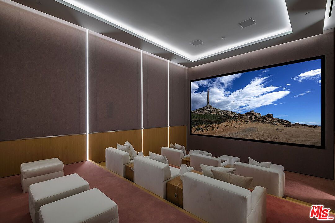 This image showcases a luxurious, modern home theater designed for an immersive cinematic experience. The room features tiered seating with plush, light-colored sofas, a large projection screen displaying a scenic landscape, and sophisticated recessed LED cove lighting that creates a sleek, ambient atmosphere. The walls are finished with acoustic paneling, emphasizing the high-end, professional-grade design of this private entertainment space.