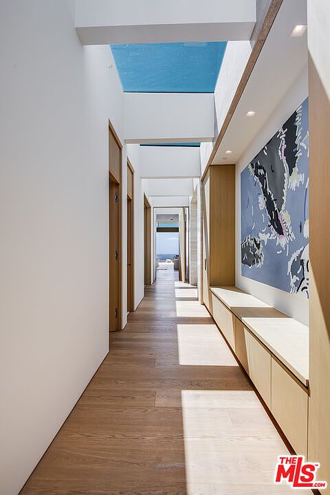 This elegant, sun-drenched hallway features a minimalist design with clean white walls, warm hardwood flooring, and a series of skylights that create a dramatic play of light and shadow. A long, custom built-in wooden console provides sleek storage, complemented by a large, abstract blue-toned art piece that adds a vibrant focal point. The perspective draws the eye down the corridor toward a bright, open space at the end, emphasizing the home's airy and sophisticated architectural flow.