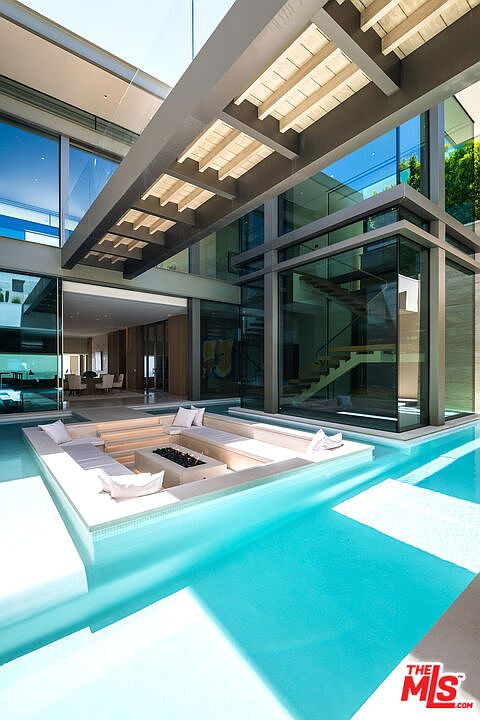 This stunning architectural masterpiece features a sunken fire pit lounge area seamlessly integrated into the center of a crystal-clear swimming pool. The modern design utilizes extensive glass walls, clean lines, and a dramatic overhead wooden pergola to create a sophisticated, resort-like atmosphere. The perspective captures the interplay between the water's reflection and the sleek, contemporary structure of the home.