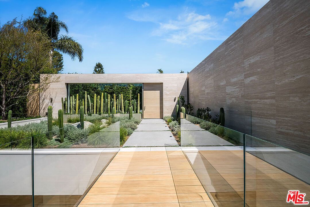 This striking modern entryway features a minimalist design with a wooden bridge walkway leading to a clean, light-toned stone facade. The entrance is framed by a lush, structured cactus garden and a large, solid wood door, creating a sophisticated and serene arrival experience. The perspective is eye-level, emphasizing the architectural symmetry and the seamless blend of natural textures with contemporary materials.