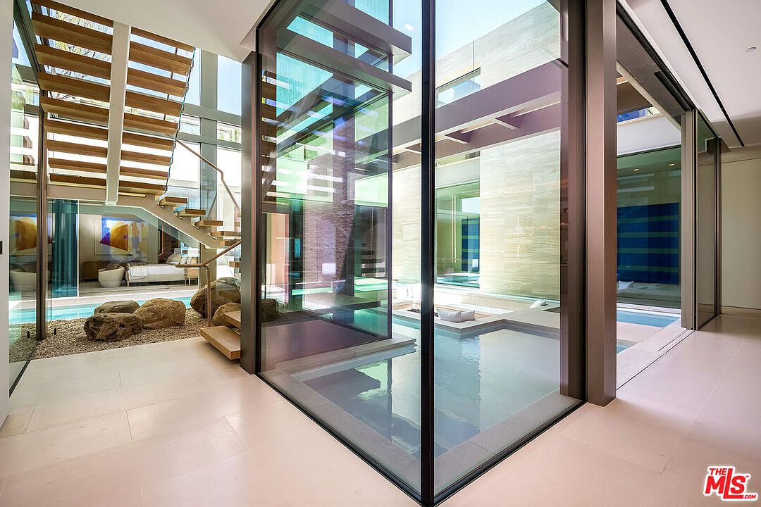This striking interior space features a dramatic floating staircase with wooden treads and glass railings, set against a backdrop of floor-to-ceiling glass walls. A central water feature with a stone-filled garden area creates a serene, architectural focal point that blurs the line between indoor and outdoor living. The design is ultra-modern and minimalist, emphasizing clean lines, natural light, and high-end materials.