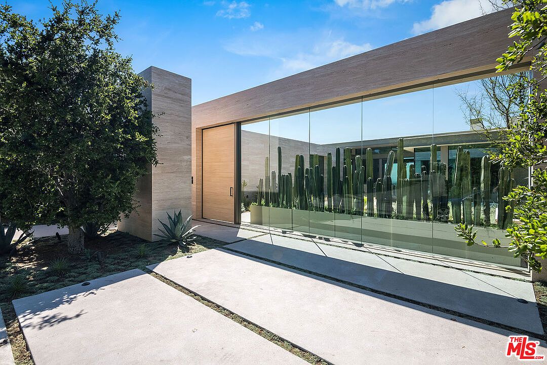This striking modern entryway features a minimalist design with clean lines, a large wooden pivot door, and expansive floor-to-ceiling glass panels that showcase an internal cactus garden. The structure is composed of light-toned stone or concrete, creating a sophisticated and architectural aesthetic. The perspective is a low-angle shot that emphasizes the grand scale of the entrance and the seamless integration of indoor and outdoor elements.