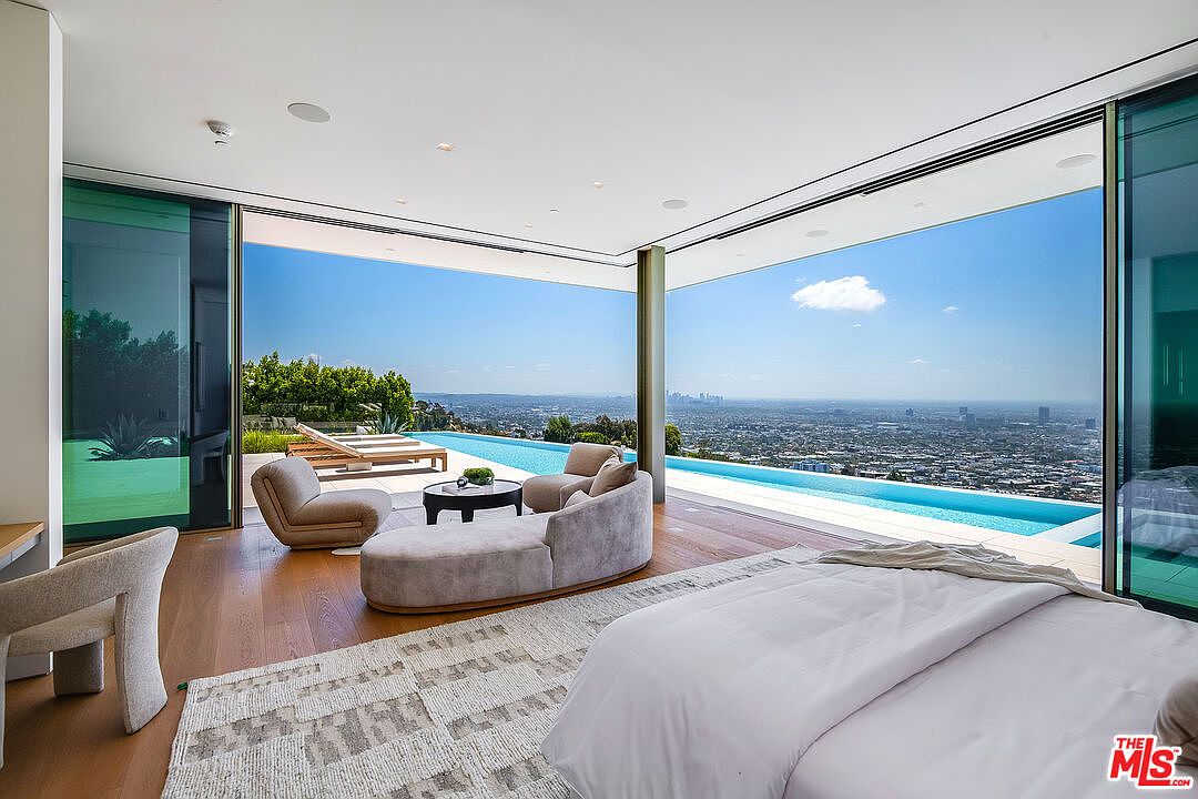 This luxurious primary bedroom features an expansive open-concept design with floor-to-ceiling glass walls that seamlessly blend the indoor space with a stunning infinity pool and panoramic city views. The room is furnished with a plush, curved sofa, modern armchairs, and a minimalist bed, all set against warm wood flooring and a textured area rug. The overall aesthetic is ultra-modern and sophisticated, emphasizing a high-end lifestyle with an emphasis on indoor-outdoor living.