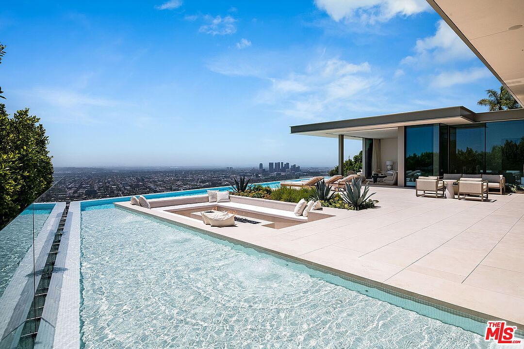 This stunning luxury estate features a sprawling infinity-edge pool that seamlessly blends into a panoramic view of the city skyline. The expansive patio includes a sunken fire pit lounge area and modern outdoor seating, all designed with a sleek, contemporary aesthetic. The bright, open space emphasizes high-end indoor-outdoor living, perfect for entertaining against a dramatic backdrop.