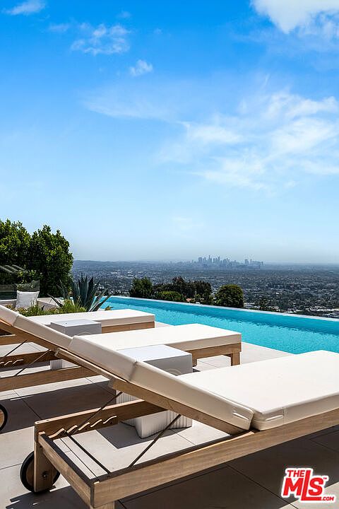 This stunning infinity-edge pool offers a breathtaking panoramic view of the distant city skyline under a clear blue sky. In the foreground, sleek wooden lounge chairs with white cushions are arranged on a modern patio, creating a luxurious and serene relaxation space. The perspective captures the seamless transition between the pool's edge and the vast horizon, emphasizing the property's high-end, resort-like atmosphere.