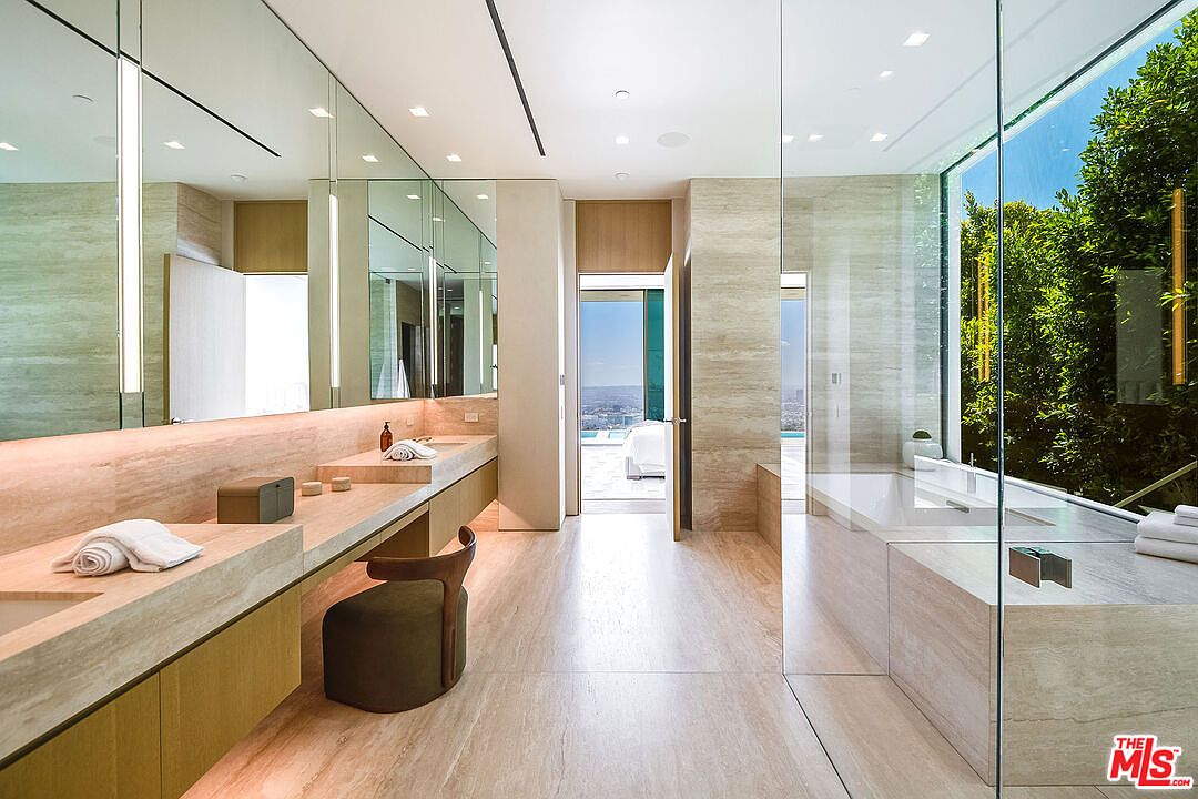 This luxurious primary bathroom features a sleek, minimalist design with warm-toned stone surfaces and expansive floor-to-ceiling mirrors that enhance the sense of space. A floating vanity with integrated lighting sits opposite a glass-enclosed soaking tub, which offers a serene view of lush greenery outside. The room flows seamlessly toward an open doorway that reveals a bright, sunlit outdoor area, creating a sophisticated and tranquil spa-like atmosphere.