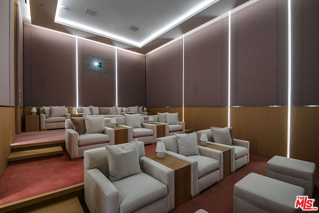 This image showcases a luxurious, tiered home theater room designed with a sophisticated, modern aesthetic. The space features multiple rows of plush, light-colored armchairs, integrated ambient wall lighting, and a projector mounted on the back wall, all set against a backdrop of rich, textured wall panels and a deep red carpet. The perspective is from the back of the room, emphasizing the tiered seating arrangement and the intimate, high-end cinematic atmosphere.
