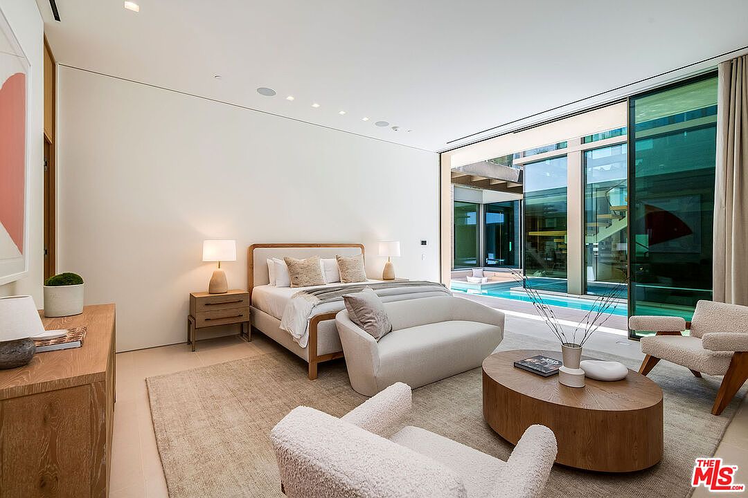This luxurious primary bedroom features a minimalist, contemporary design with a neutral color palette and high-end finishes. The room is anchored by a plush bed and a curved sofa, leading directly to a floor-to-ceiling glass wall that opens onto a private pool area. The seamless transition between the indoor living space and the outdoor water feature creates a serene, resort-like atmosphere.
