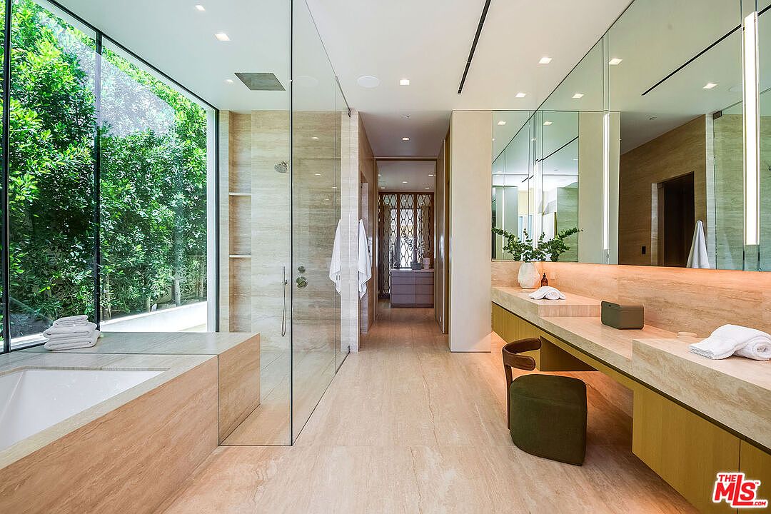 This luxurious primary bathroom features a seamless blend of natural stone surfaces, a large glass-enclosed walk-in shower, and a deep soaking tub positioned next to floor-to-ceiling windows that overlook lush greenery. A long, floating vanity with integrated lighting and a plush stool creates a sophisticated spa-like atmosphere. The perspective is centered, drawing the eye through the elegant space toward a secondary room in the background, emphasizing the open and airy architectural design.