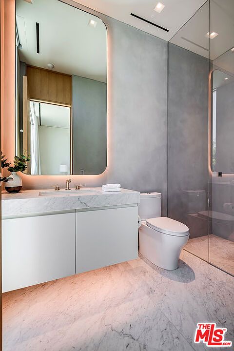 This sleek, modern primary bathroom features a floating white vanity topped with elegant marble, complemented by a large, backlit rounded mirror. The space is finished with soft gray textured walls and light marble flooring, creating a serene and luxurious atmosphere. A glass-enclosed shower and a minimalist toilet complete the clean, contemporary design.