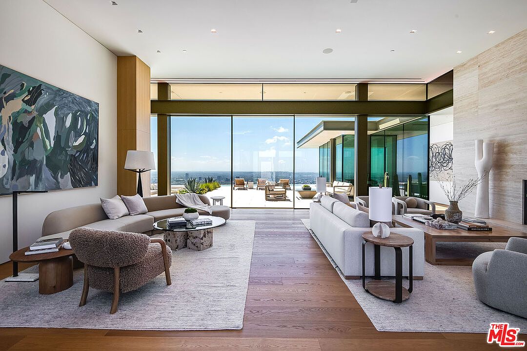 This expansive, modern living room features a sophisticated open-concept design with floor-to-ceiling glass walls that frame a breathtaking panoramic city view. The space is elegantly furnished with two large, neutral-toned sectional sofas, textured accent chairs, and a central stone coffee table, all resting on plush area rugs atop warm hardwood flooring. The room exudes a luxurious, airy atmosphere, seamlessly blending high-end interior design with the outdoor terrace and horizon beyond.