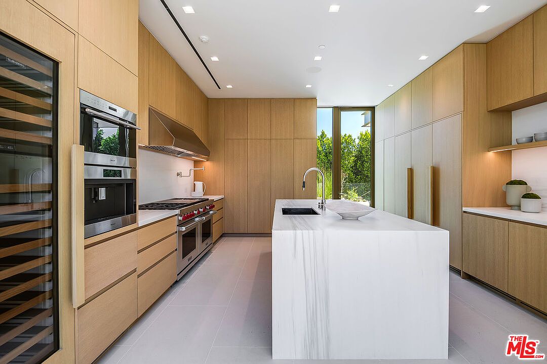This sleek, modern kitchen features light wood cabinetry, a large waterfall-edge marble island, and high-end stainless steel appliances, including a built-in wine refrigerator. The minimalist design is complemented by recessed lighting and a seamless transition to the outdoors through a floor-to-ceiling window. The overall aesthetic is sophisticated, clean, and highly functional, perfect for contemporary living.