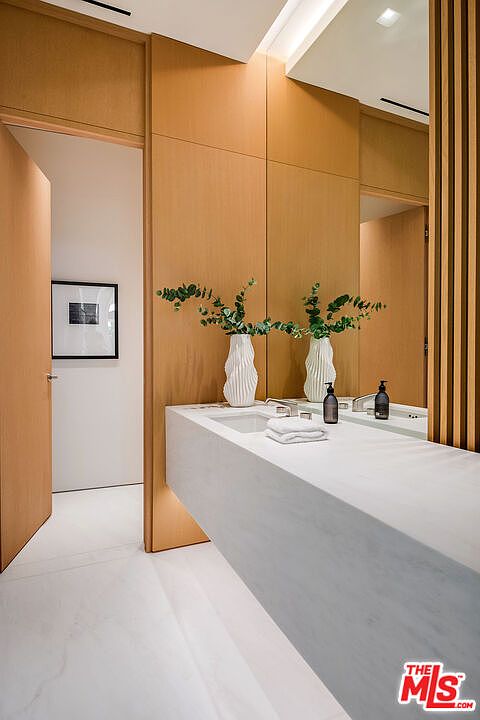 This sleek, modern bathroom features a floating white marble vanity set against warm, wood-paneled walls that create a sophisticated and serene atmosphere. Two sculptural white vases filled with greenery sit atop the counter, reflecting in the expansive mirror, while a minimalist doorway leads to an adjacent space. The perspective is captured from a slightly elevated angle, emphasizing the clean lines and high-end materials of this contemporary design.