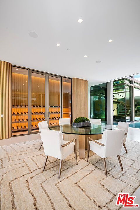This sophisticated dining area features a circular glass-top table surrounded by four modern white upholstered chairs, set atop a textured, geometric-patterned area rug. The space is anchored by a sleek, floor-to-ceiling glass-enclosed wine cellar with warm interior lighting, creating a luxurious and inviting atmosphere. Large windows provide a seamless connection to the outdoor pool area, enhancing the open-concept, contemporary design of the home.