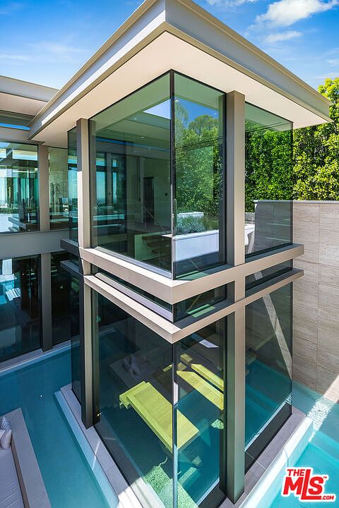 This striking architectural shot captures a modern, multi-level glass structure that appears to float directly within a serene, turquoise swimming pool. The design emphasizes clean lines and transparency, with the reflection of the surrounding greenery on the glass panels creating a seamless blend between the home and nature. The perspective from above highlights the unique integration of water and contemporary living space, showcasing a luxurious and sophisticated aesthetic.