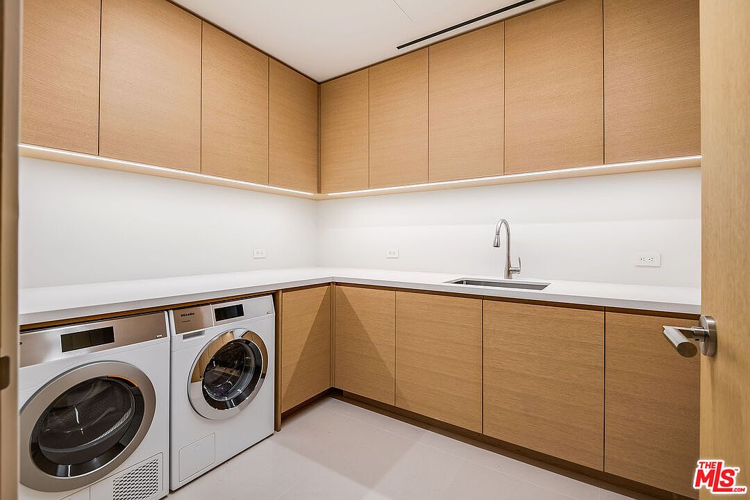 This modern, minimalist laundry room features sleek light wood cabinetry, a crisp white countertop, and a built-in stainless steel sink. The space is equipped with high-end Miele washer and dryer units, creating a clean and highly functional utility area. The under-cabinet lighting adds a sophisticated touch to the bright, contemporary design.