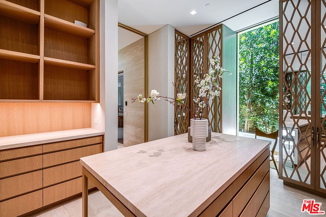 This sophisticated walk-in closet features custom light wood cabinetry, a central island with a stone countertop, and elegant lattice-patterned room dividers. The space is bathed in natural light from a large window overlooking lush greenery, creating a serene and luxurious atmosphere. The clean lines and high-end materials suggest a modern, high-end residential design.