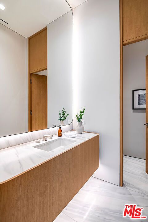 This modern, minimalist bathroom features a sleek wood-paneled vanity topped with a white marble countertop and an integrated sink. A large, frameless arched mirror reflects the clean lines of the space, while a small vase with greenery adds a touch of organic warmth. The room is finished with light-colored marble flooring and a doorway leading to an adjacent space, creating a bright and sophisticated atmosphere.