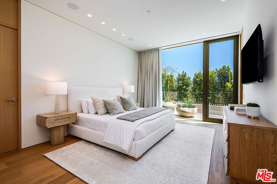 This elegant primary bedroom features a minimalist, contemporary design with a plush white bed as the centerpiece, complemented by light wood nightstands and a matching dresser. Large floor-to-ceiling glass doors open onto a private balcony, flooding the room with natural light and offering a serene view of lush greenery. The neutral color palette and clean lines create a sophisticated, tranquil atmosphere perfect for a luxury residence.
