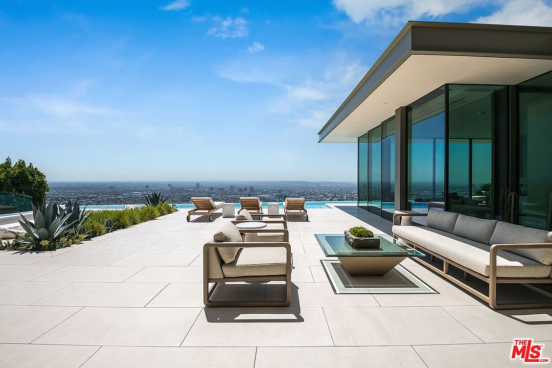 This expansive, modern outdoor terrace features a sleek infinity-edge pool overlooking a panoramic city skyline. The space is furnished with contemporary lounge seating, including a long sofa and armchairs, arranged on light-colored stone pavers. The architectural design emphasizes clean lines and seamless indoor-outdoor flow through floor-to-ceiling glass walls.
