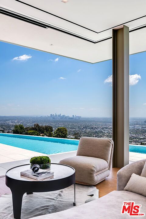This sophisticated indoor-outdoor living space features a seamless transition to a stunning infinity pool overlooking a panoramic city skyline. The interior is furnished with plush, neutral-toned seating and a minimalist circular coffee table, emphasizing a modern, luxurious aesthetic. The expansive glass walls and open-concept design create an airy, cinematic atmosphere that highlights the breathtaking elevated view.