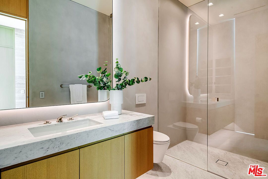 This modern, minimalist bathroom features a sleek marble-topped vanity with a light wood cabinet and a large, frameless mirror. A glass-enclosed walk-in shower with a soft, integrated light strip creates a spa-like atmosphere, complemented by clean lines and neutral tones. The perspective is a straight-on shot that highlights the high-end finishes and sophisticated, contemporary design of the space.