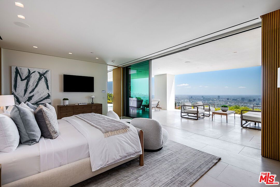 This luxurious primary bedroom features a seamless transition to an expansive outdoor terrace through floor-to-ceiling glass sliding doors, offering a breathtaking panoramic view of the city skyline. The room is decorated in a sophisticated, neutral palette with a plush bed, textured area rug, and modern minimalist furniture. The open-concept design emphasizes a high-end, airy atmosphere that blends indoor comfort with stunning natural vistas.