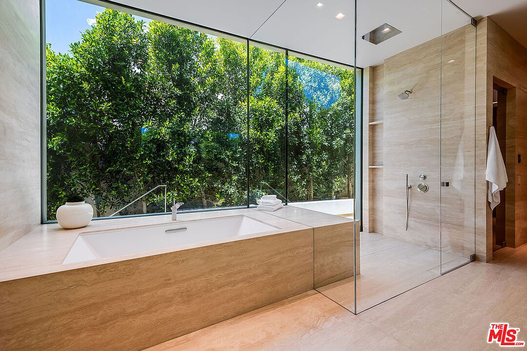 This luxurious primary bathroom features a sleek, minimalist design with floor-to-ceiling glass walls that offer an immersive view of lush greenery outside. The space includes a deep, built-in soaking tub encased in light-toned stone and a spacious walk-in shower area separated by a frameless glass partition. The neutral color palette and high-end materials create a serene, spa-like atmosphere perfect for relaxation.