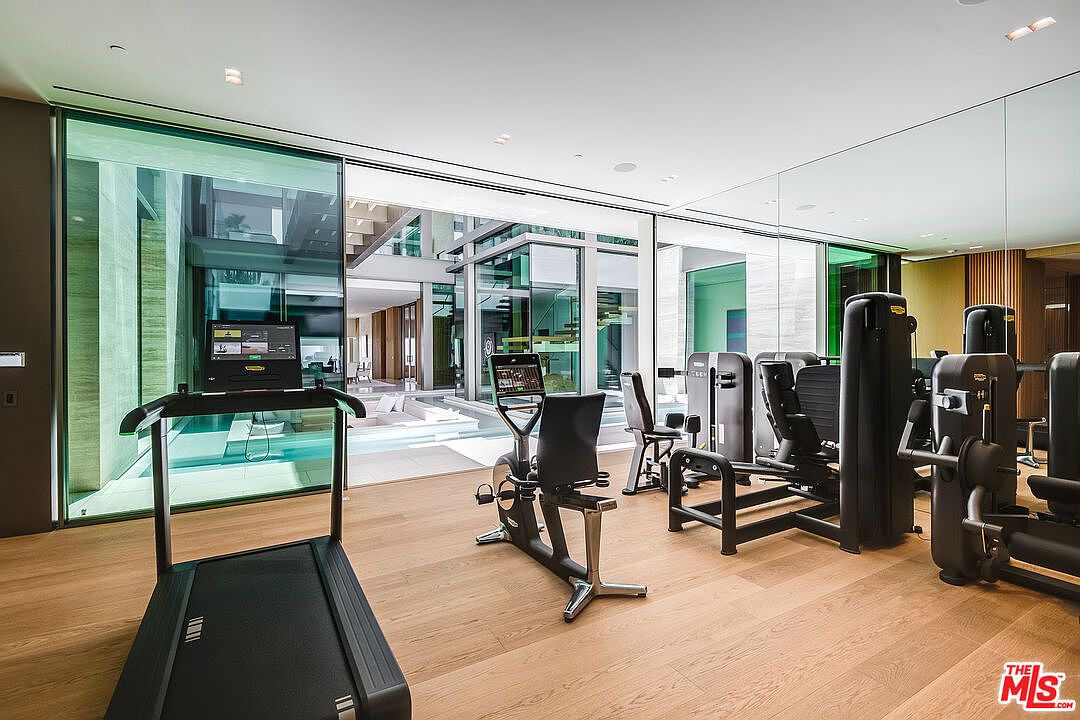 This sleek, modern home gym features high-end Technogym equipment, including a treadmill and a stationary bike, set against a backdrop of floor-to-ceiling glass walls. The room offers a seamless indoor-outdoor connection, looking out onto a serene courtyard with a pool, creating an airy and luxurious workout environment. The light wood flooring and minimalist design emphasize a sophisticated, high-end aesthetic.