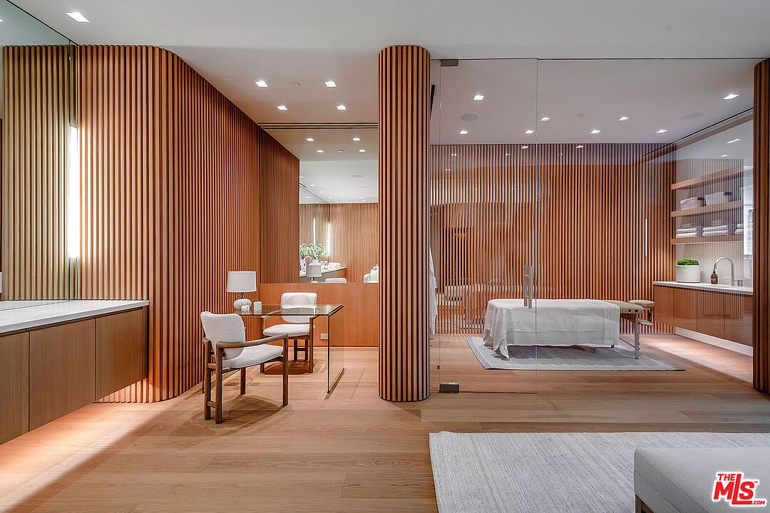 This sophisticated home spa or wellness room features warm, vertical wood slat paneling that creates a serene and luxurious atmosphere. The space includes a glass-enclosed massage area with a treatment table, a minimalist desk setup, and recessed lighting throughout. The combination of natural wood tones and clean, modern lines provides a high-end, tranquil aesthetic perfect for relaxation.