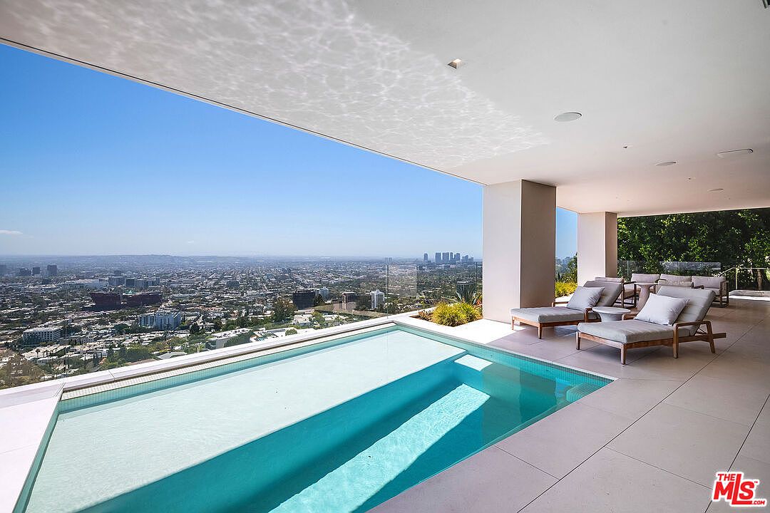 This stunning outdoor terrace features a sleek, infinity-edge swimming pool that seamlessly blends into a panoramic, high-angle view of the sprawling city skyline below. The space is furnished with modern, minimalist lounge chairs arranged under a covered patio, creating a luxurious and serene atmosphere. The clean architectural lines and expansive open-air design emphasize a sophisticated, high-end lifestyle.