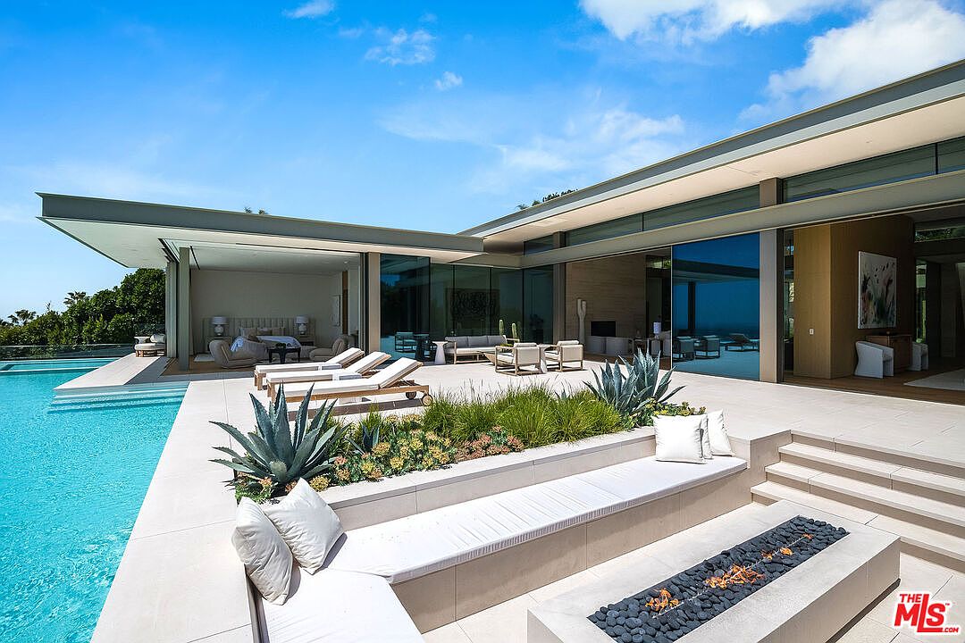 This stunning modern luxury estate features a seamless indoor-outdoor flow with expansive glass walls opening onto a pristine pool deck. The area includes a sunken fire pit lounge, lush desert-inspired landscaping, and sleek lounge chairs overlooking a sparkling infinity-edge pool. The architectural design emphasizes clean lines, neutral stone textures, and a sophisticated, resort-like atmosphere.