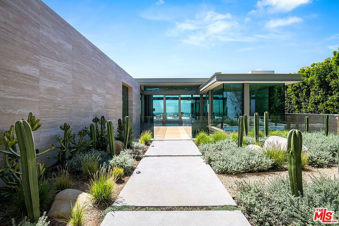 This striking modern entryway features a clean, minimalist aesthetic with a long concrete walkway leading toward a glass-walled foyer. The structure is accented by a large, textured stone wall on the left and lush desert landscaping, including various cacti and native shrubs, that frames the path. The perspective is a low-angle, eye-level shot that emphasizes the architectural symmetry and the seamless transition between the outdoor garden and the interior living space.