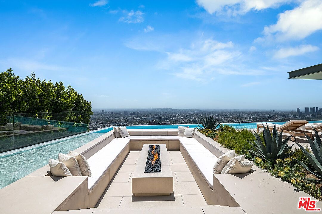 This stunning outdoor space features a sunken fire pit lounge area integrated directly into a sleek infinity-edge pool, offering panoramic views of the city skyline. The modern design utilizes clean lines, light-toned stone, and minimalist landscaping, creating a luxurious and serene atmosphere. The perspective captures the seamless transition between the architectural seating area and the expansive horizon beyond.