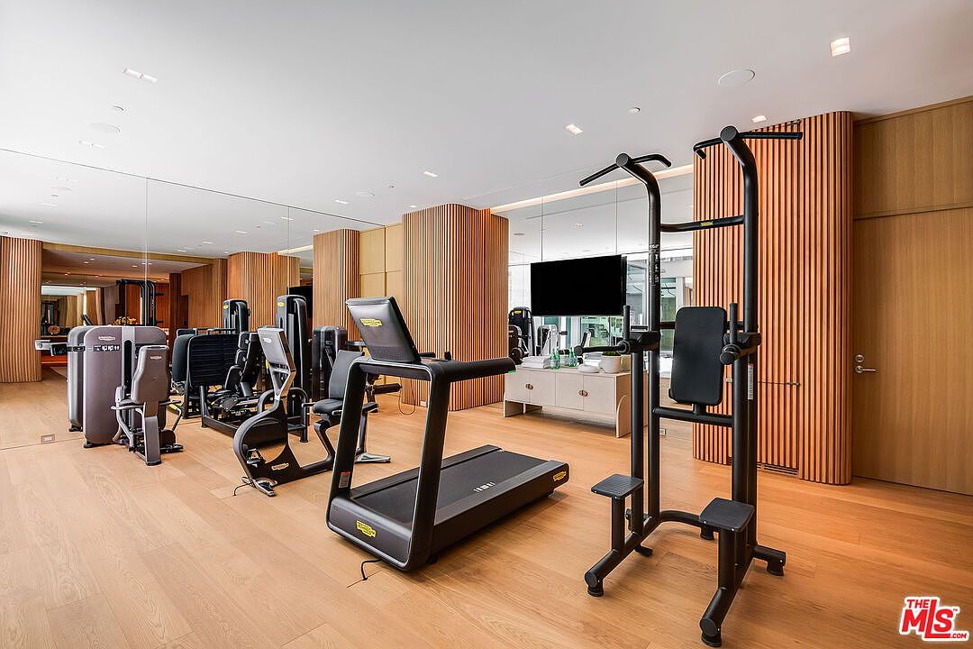 This high-end home gym features a sleek, modern aesthetic with light wood flooring and warm, vertical wood-slatted wall paneling. The space is equipped with professional-grade Technogym equipment, including a treadmill, strength training machines, and a pull-up/dip station, all reflected in a large wall-to-wall mirror. A flat-screen television and a minimalist console table add a touch of luxury, creating a sophisticated and functional workout environment.