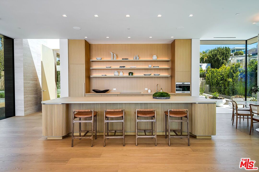 This sophisticated, modern kitchen features a large, fluted-wood island with four woven-seat bar stools, serving as the centerpiece of the room. The back wall is composed of light oak cabinetry with integrated shelving and recessed lighting, creating a warm and minimalist aesthetic. Large floor-to-ceiling windows on the right provide a seamless transition to the outdoor greenery, bathing the space in natural light.