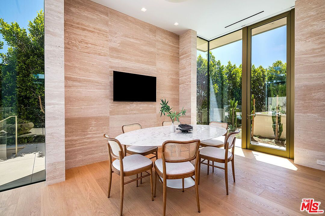 This elegant dining room features a minimalist aesthetic with a light-toned wood floor and a striking floor-to-ceiling travertine accent wall. An oval marble-topped table is surrounded by six mid-century modern cane-back chairs, while expansive glass walls provide a seamless connection to the lush outdoor greenery. The space is bright and airy, offering a sophisticated atmosphere perfect for refined entertaining.