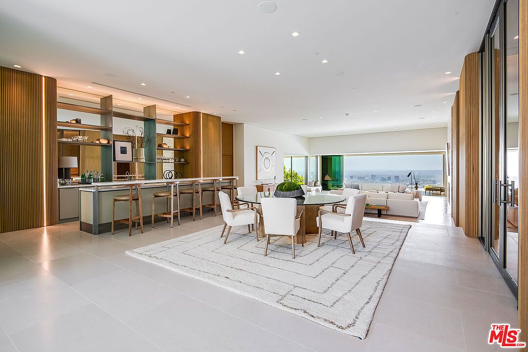 This expansive, open-concept living and dining area exudes modern luxury with its minimalist aesthetic, high ceilings, and floor-to-ceiling windows that frame a breathtaking panoramic city view. The space features a sophisticated bar area with integrated lighting, a circular dining table set on a textured area rug, and a plush seating arrangement in the background. The warm wood paneling and neutral color palette create a serene, high-end atmosphere perfect for entertaining.