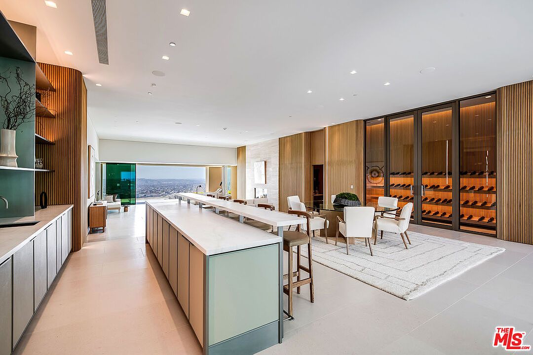 This expansive, ultra-modern kitchen features a long, sleek island with a white countertop and light green cabinetry, complemented by a minimalist design aesthetic. To the right, a sophisticated glass-enclosed wine cellar serves as a focal point, while the open-concept layout flows seamlessly toward a dining area and a floor-to-ceiling glass wall revealing a stunning panoramic city view. The space is defined by warm wood paneling, recessed lighting, and a clean, high-end contemporary finish.