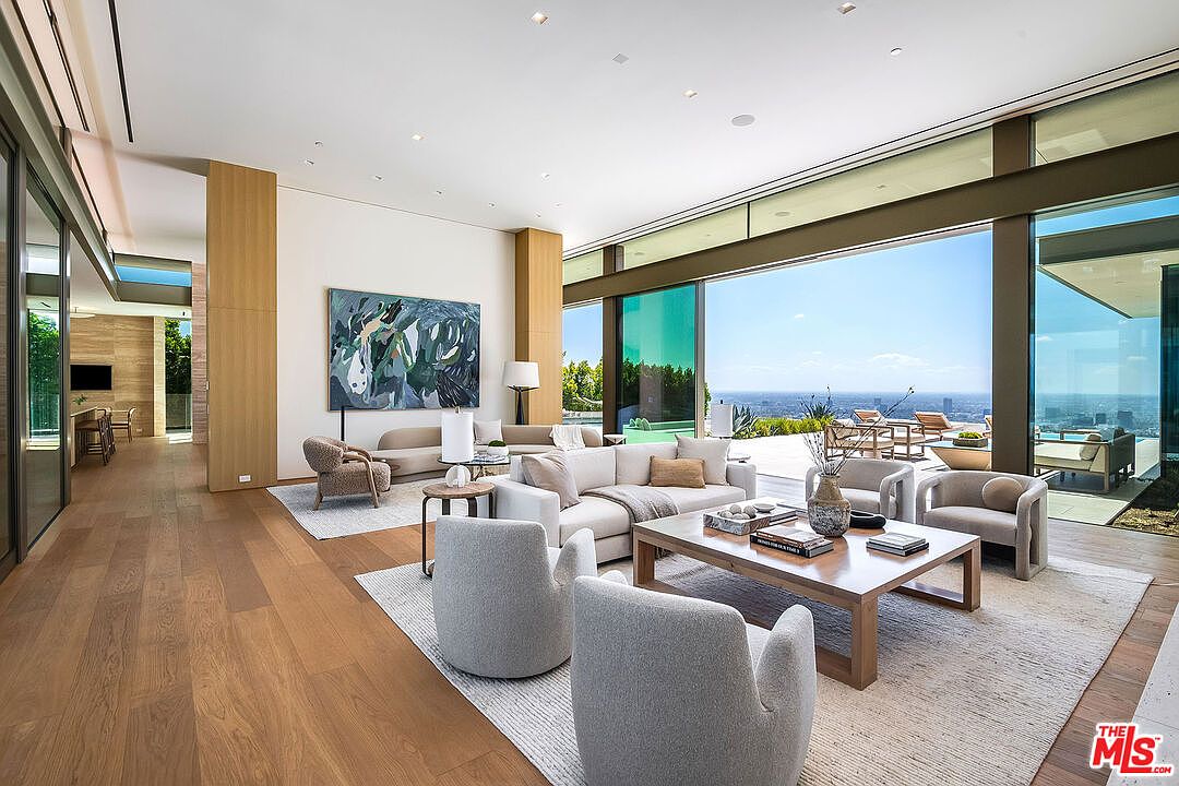 This expansive, modern living room features an open-concept design with floor-to-ceiling glass walls that seamlessly blend the interior with a stunning panoramic city view. The space is furnished with plush, neutral-toned seating, a large wooden coffee table, and a striking abstract painting, all set against warm hardwood floors and high ceilings. The overall aesthetic is one of sophisticated luxury, emphasizing light, space, and a connection to the outdoors.