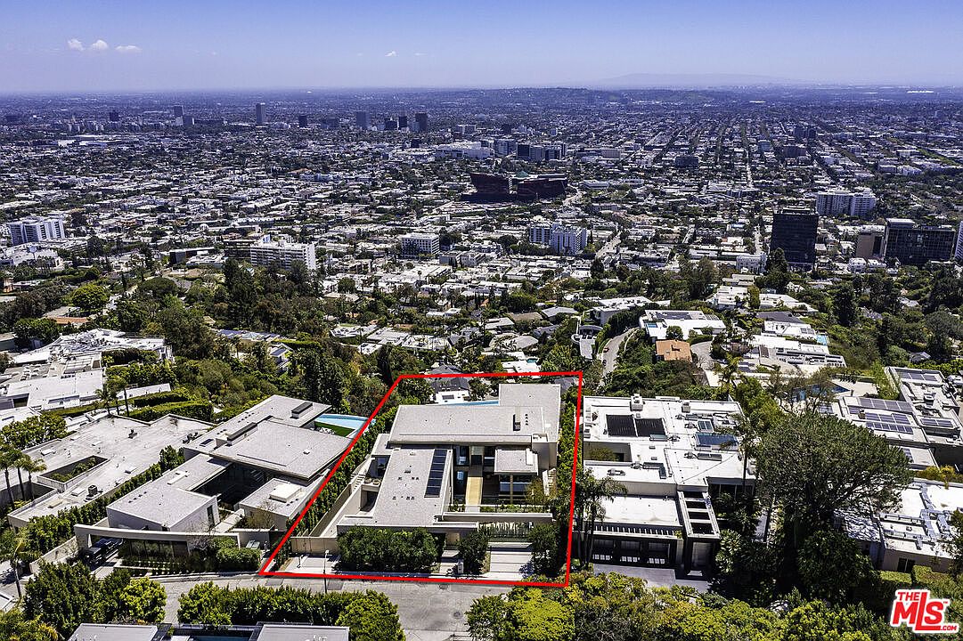 This high-angle aerial shot captures a modern, multi-level luxury residence nestled in a lush, hilly neighborhood with a sprawling cityscape in the background. The property, highlighted by a red outline, features a flat-roof architectural design, a private swimming pool, and well-manicured landscaping. The perspective emphasizes the home's prime location and its integration into the dense, upscale urban environment.