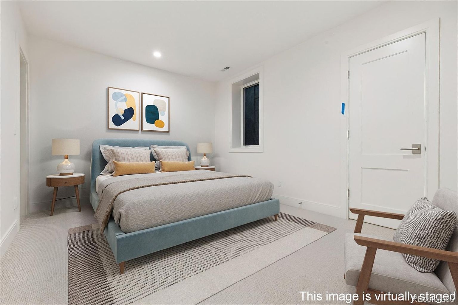 This is a staged interior shot of a bedroom. The room features a bed with a light blue frame, neutral bedding, and decorative pillows. Two framed abstract art pieces hang above the bed, and there are side tables with lamps on either side. A modern armchair sits in the corner, and a striped rug partially covers the carpeted floor. A window and a white door are also visible.