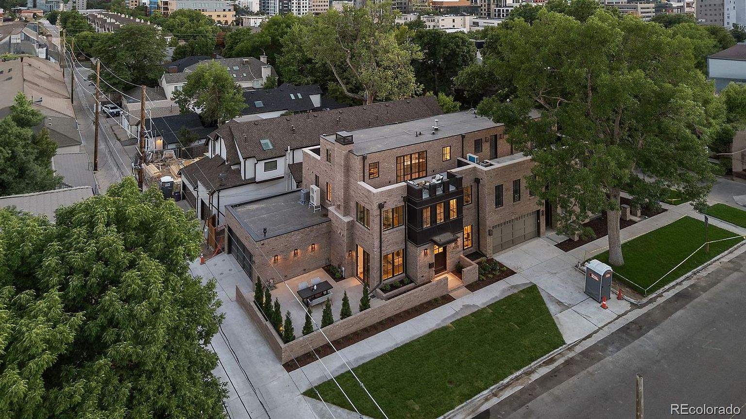 This aerial shot showcases a modern brick home with a flat roof and a well-manicured lawn. The property features a private patio area with outdoor seating, a two-car garage, and mature trees providing shade. The overall impression is one of sophisticated urban living.