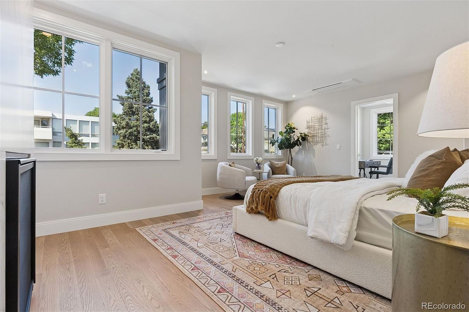 This is a bright and airy primary bedroom featuring a large bed with a white textured base and white linens, complemented by a patterned area rug. The room is well-lit by several windows offering natural light, and includes a seating area with two chairs and a small table. The overall impression is one of comfort and sophistication.