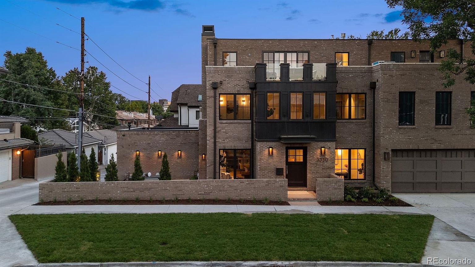 This is a front exterior view of a modern brick townhouse. The house features a combination of light brown brick and dark metal accents, with large windows and a small balcony on the upper level. A well-manicured lawn and a low brick wall add to the property's curb appeal.