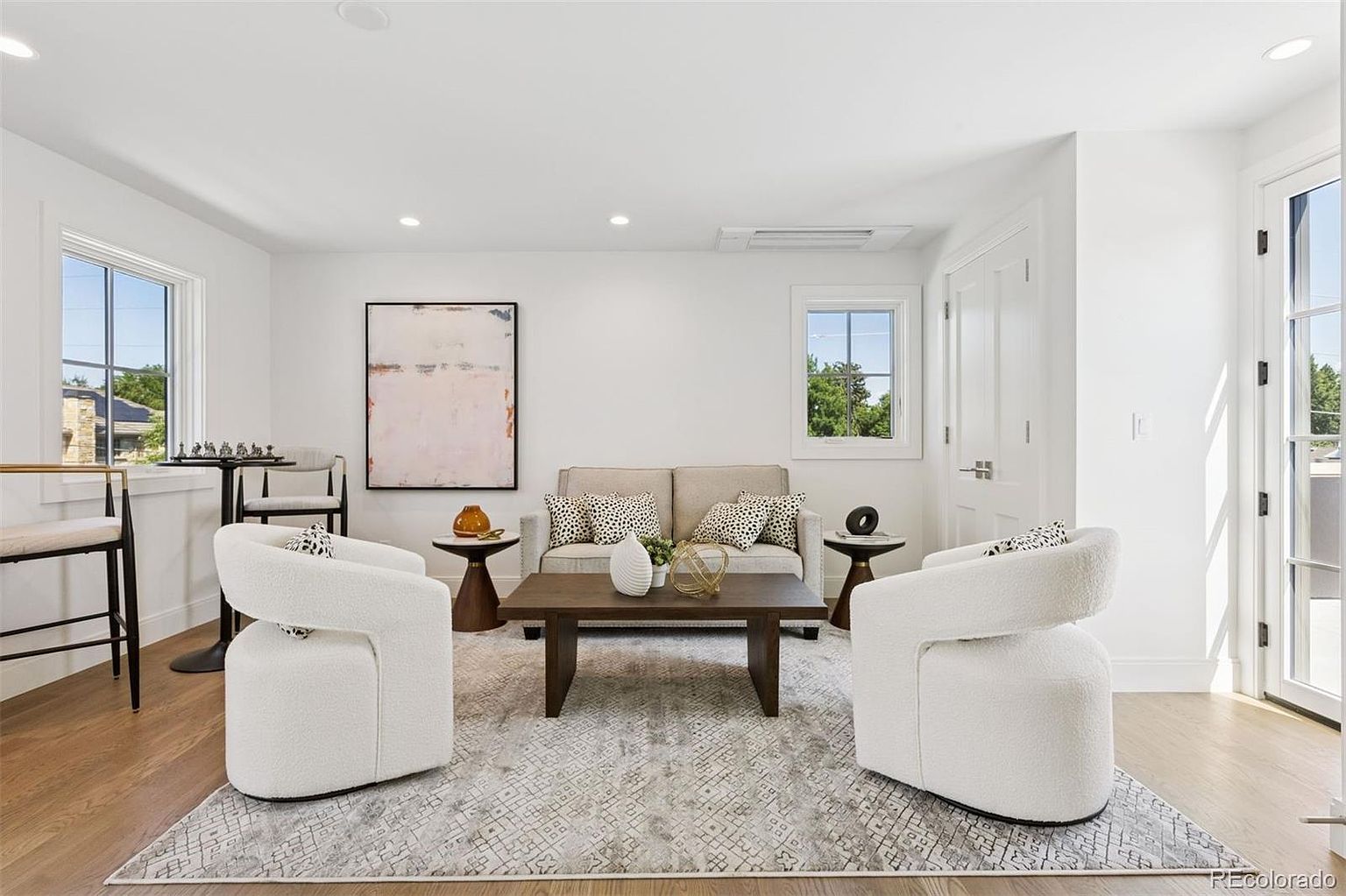 This is a bright and airy living room featuring a neutral color palette with white walls and light wood flooring. The room is furnished with a beige sofa, two white armchairs, and a dark wood coffee table, all arranged on a patterned area rug. Natural light streams in through the windows, creating a welcoming and comfortable atmosphere.