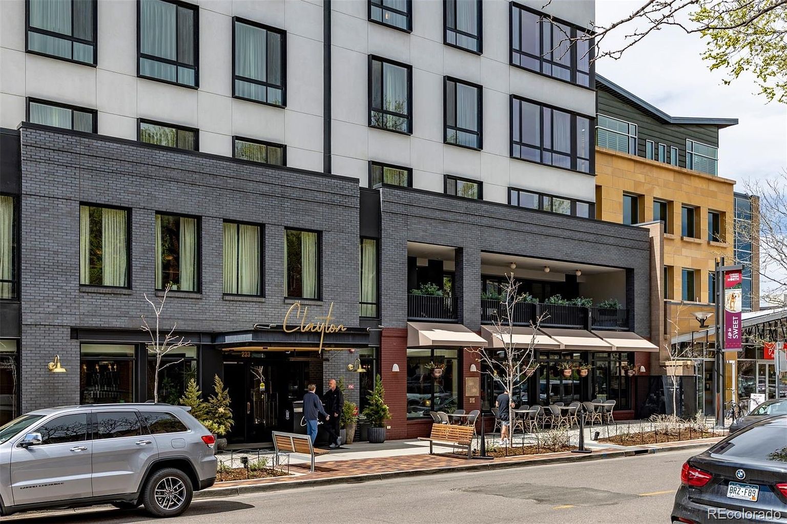 The image showcases the front exterior of a modern building, likely a hotel or mixed-use development, featuring a combination of gray brick and white facade elements. The building has large windows and a prominent entrance with the name "Clayton" displayed above. Outdoor seating and landscaping add to the inviting street-level presence, with cars parked on the street indicating an urban setting.