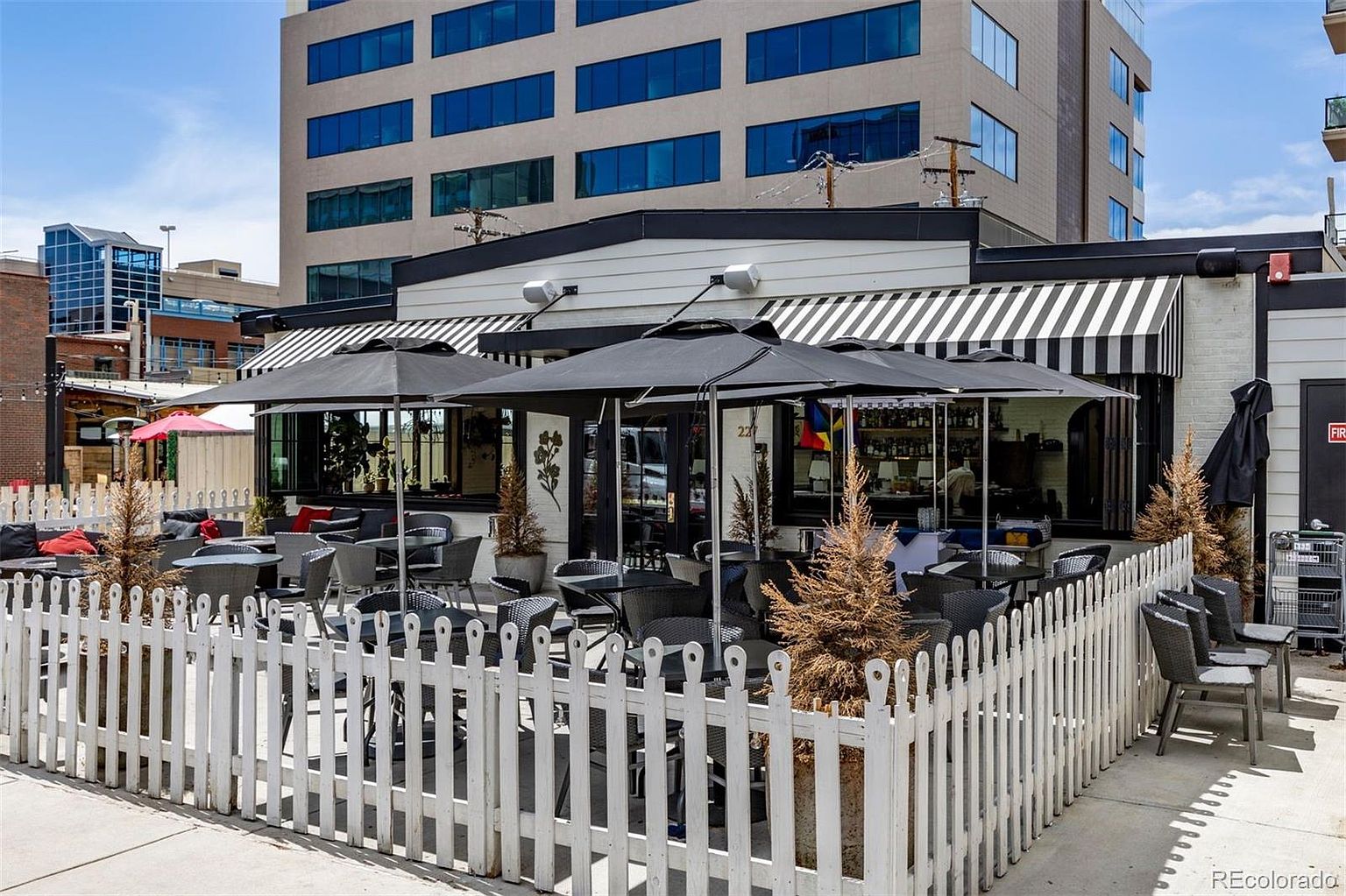 This image showcases an inviting outdoor patio area, likely belonging to a restaurant or cafe. The space is enclosed by a white picket fence and features numerous tables and chairs under large black umbrellas, providing shade. The building behind the patio has a black and white striped awning and a modern architectural style, suggesting a sophisticated and comfortable dining experience.