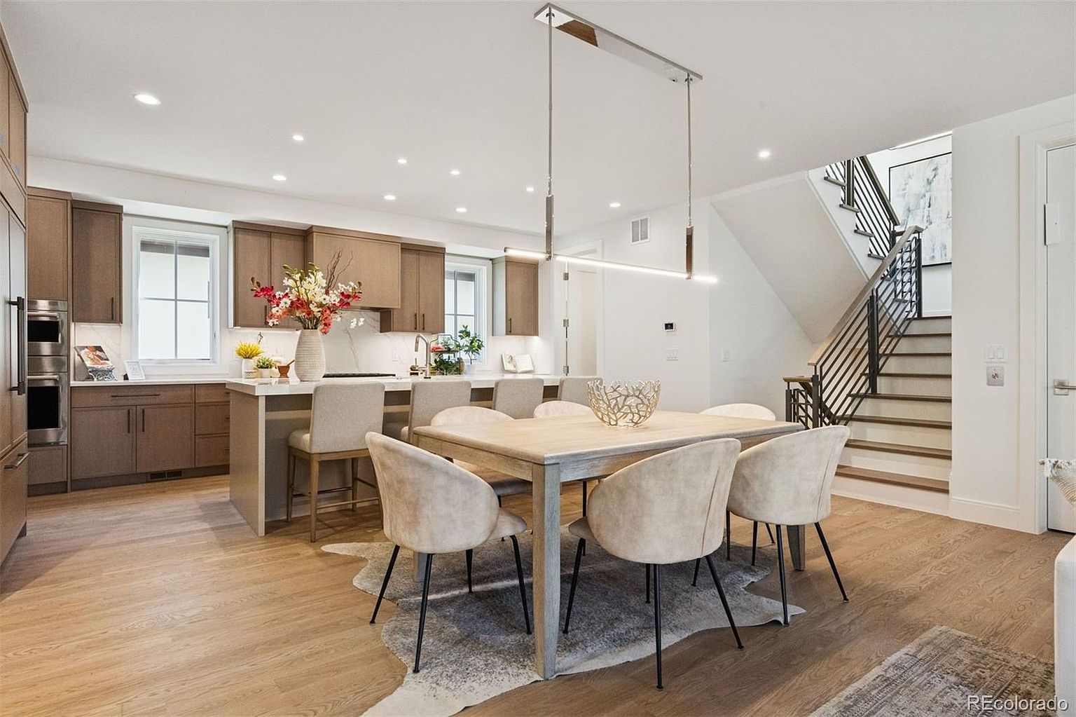 This interior shot showcases a dining area adjacent to a modern kitchen. The dining space features a light wood table surrounded by six upholstered chairs with black legs, set upon a cowhide rug. A contemporary linear chandelier hangs above the table, and the open layout leads into a kitchen with sleek cabinetry and a staircase with metal railings, creating a bright and inviting atmosphere.