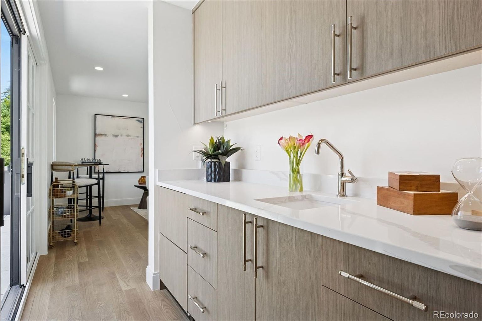 This interior shot showcases a modern kitchen area with sleek, light wood-grain cabinets and drawers, complemented by a white countertop and backsplash. A stainless steel faucet and sink are visible, along with decorative items like flowers and wooden boxes. The space opens to a bright hallway and dining area, creating an inviting and functional atmosphere.